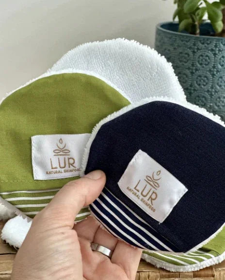 Hand met herbruikbare katoenen gezichtsreinigingspads in groen en donkerblauw, met een plant op de achtergrond.